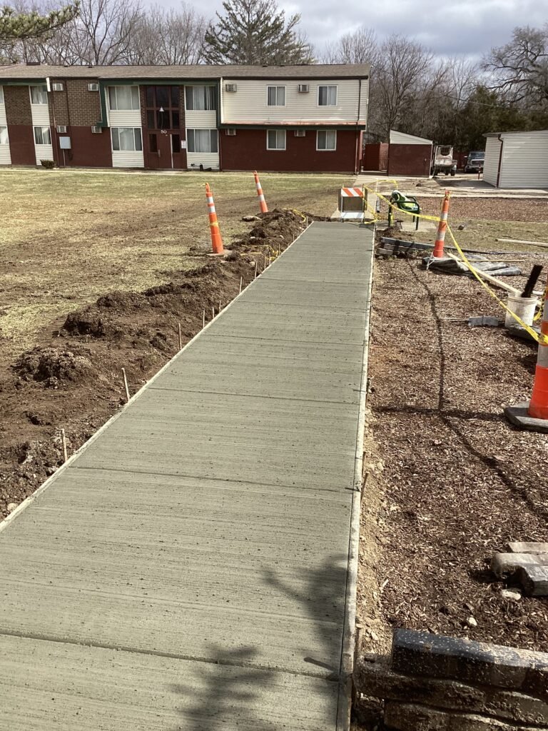 A newly poured concrete sidewalk by Empire Paving stretches past orange traffic cones and caution tape, beside a grassy area in front of an apartment building on a cloudy day.