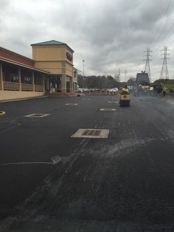 Workers from Empire Paving are paving a parking lot outside a commercial building on a cloudy day. Construction equipment and vehicles are visible, with steam rising from the freshly laid asphalt.
