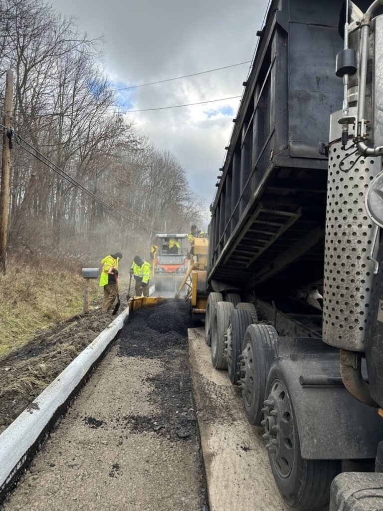 Construction workers in safety vests repave a rural road using heavy machinery from a commercial asphalt company, with piles of asphalt next to a grassy embankment and bare trees under a cloudy sky.