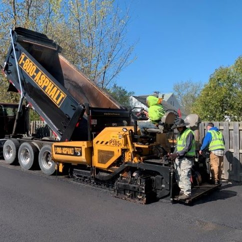 Construction workers from Empire Paving operate an asphalt paving machine, laying new asphalt from a dump truck with “1-800-ASP-HALT” on its side, on a sunny day with trees and a fence in the background.