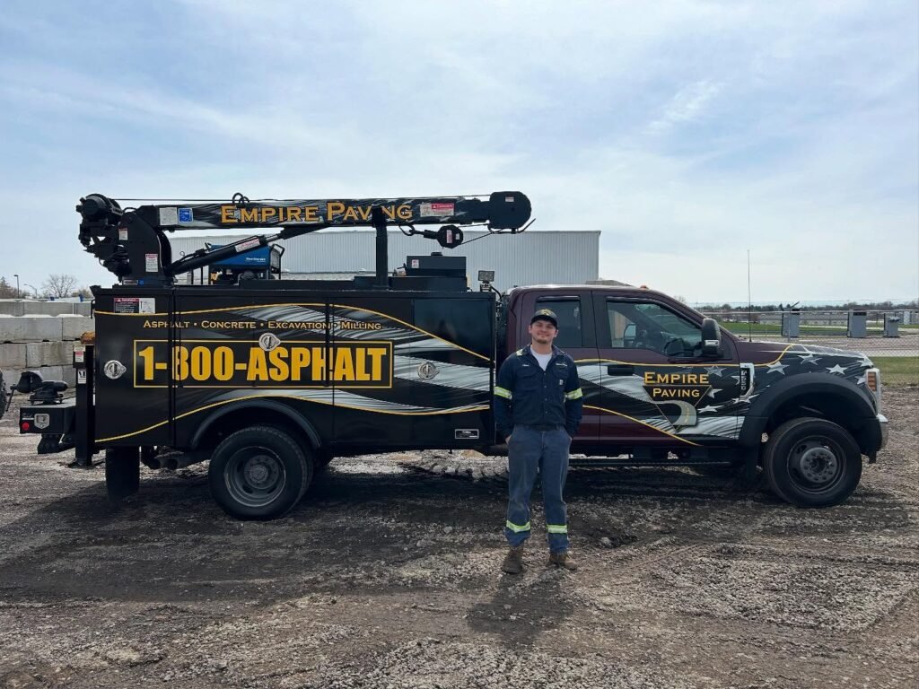A worker in uniform stands before a black Empire Paving truck with a mounted crane. As one of the top asphalt companies Cleveland Ohio trusts, they offer commercial concrete paving contractor services and display 1-800-ASPHALT.