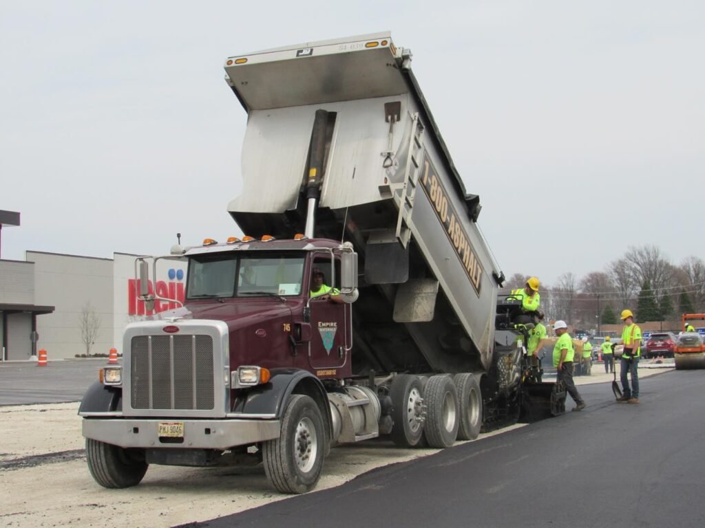 A dump truck unloads asphalt into a paving machine as several construction workers in safety gear, part of a leading commercial asphalt company near me, guide the process and work on freshly laid pavement by the roadside.