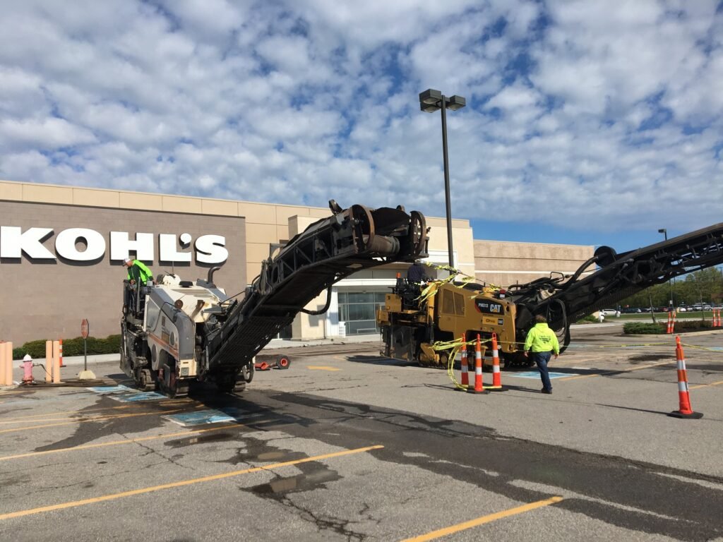 Workers from an Ohio paving and construction crew operate large road machinery in a shopping center parking lot near Kohl’s, where orange cones and barriers mark the work zone under a partly cloudy sky.