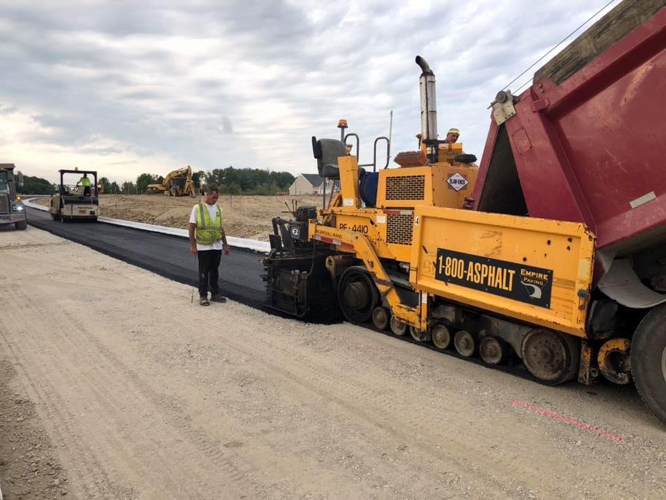 An Empire Paving yellow asphalt paver machine lays new black asphalt on a road under construction, with a worker in a safety vest nearby and more construction vehicles in the background under an overcast sky.