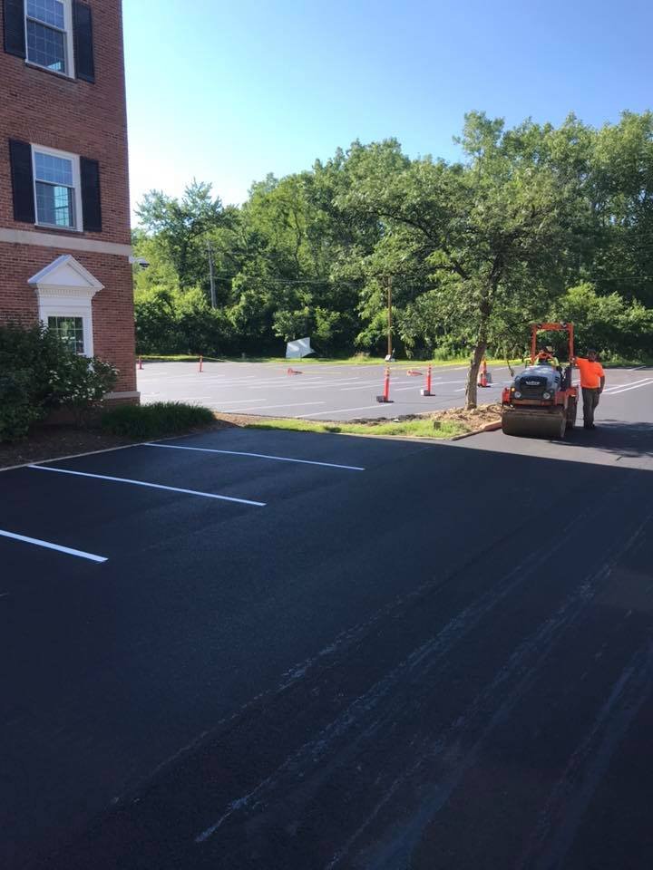 Freshly paved parking lot with white lines, bordered by a red brick building on the left. Two workers from an Ohio paving and construction crew operate machinery near an orange cone and a small tree. Trees and traffic cones are in the background.