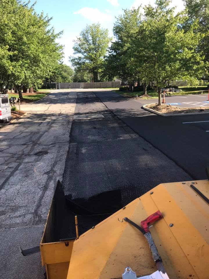 View from construction vehicle of a road being resurfaced by Empire Paving; the central section is stripped and ready for paving, with fresh asphalt on one side. Trees and parked cars line the road under a clear sky.