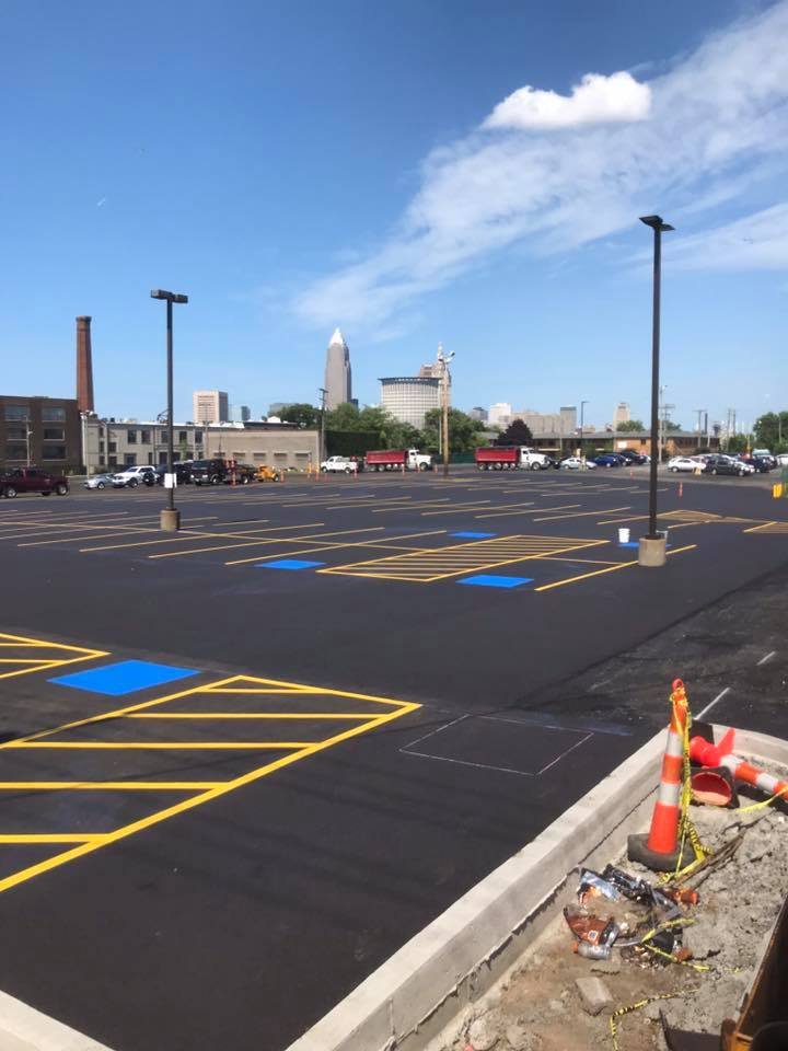 A newly paved parking lot by Ohio Paving and Construction features blue accessible spaces, yellow lines, and light poles. Traffic cones and construction materials are in the foreground, all set against a city skyline under a bright blue sky.