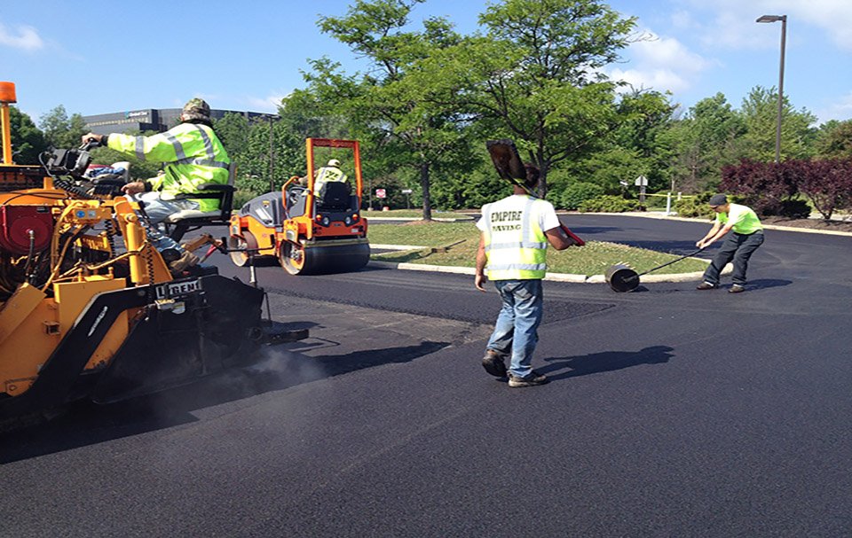 Workers in safety vests operate heavy machinery and smooth fresh asphalt while paving a road on a sunny day, with green trees and blue sky in the background—another quality job by a top commercial concrete paving contractor.