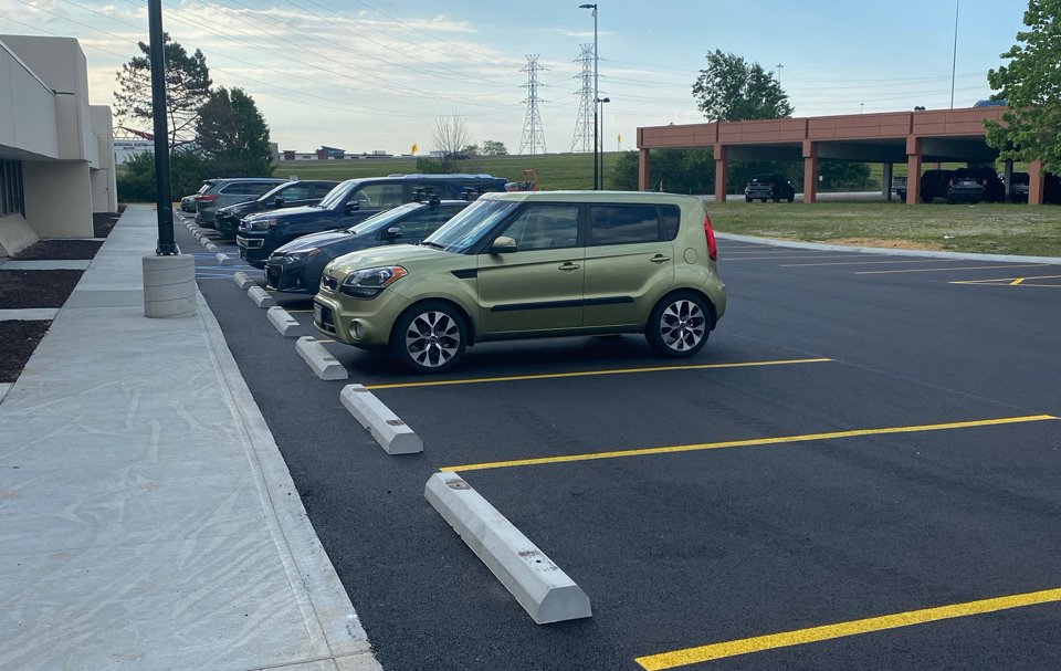 A light green compact car is parked unevenly across two parking spaces in a nearly empty lot, recently resurfaced by empire paving. Several other cars are parked correctly nearby, with trees and a building visible in the background.