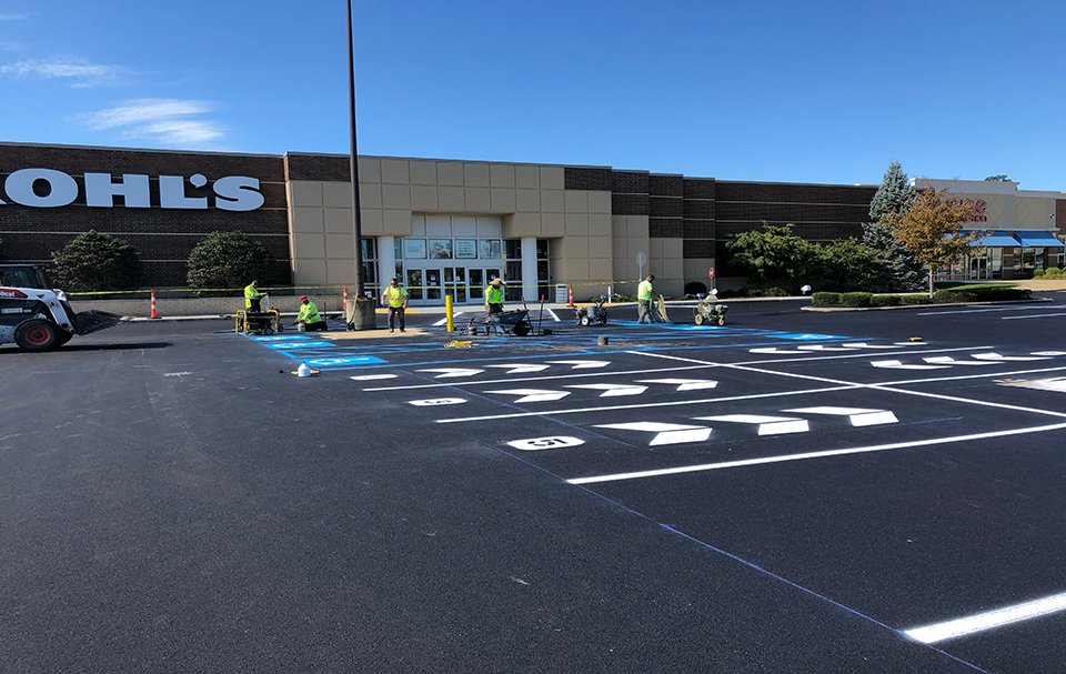 Workers in safety vests paint lines and arrows on a freshly paved parking lot in front of a Kohl’s store, completed by one of the top commercial asphalt companies near me, with clear blue sky and landscaping visible in the background.