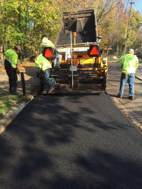 Workers in bright safety vests operate a paving machine laying fresh asphalt on a road, with trees and fallen leaves in the background—an example of quality work by a commercial concrete paving contractor.