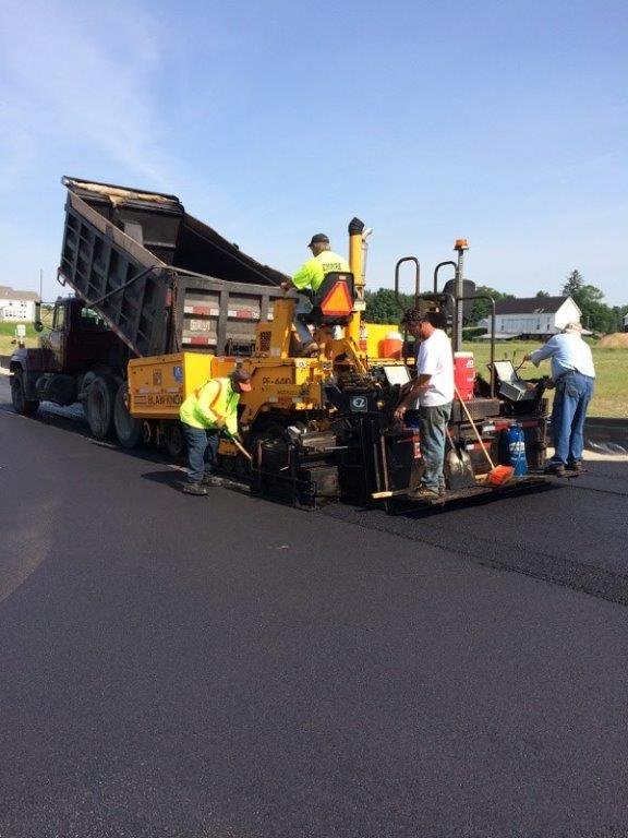 Workers operate a paving machine laying fresh asphalt on a road, while a dump truck unloads hot asphalt into the machine—showcasing the expertise of one of the top asphalt companies Cleveland Ohio relies on, all under sunny, clear skies.