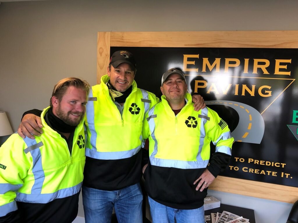 Three men wearing bright yellow reflective jackets smile and pose together in front of an Empire Paving sign inside an office, representing the team behind Ohio paving and construction.