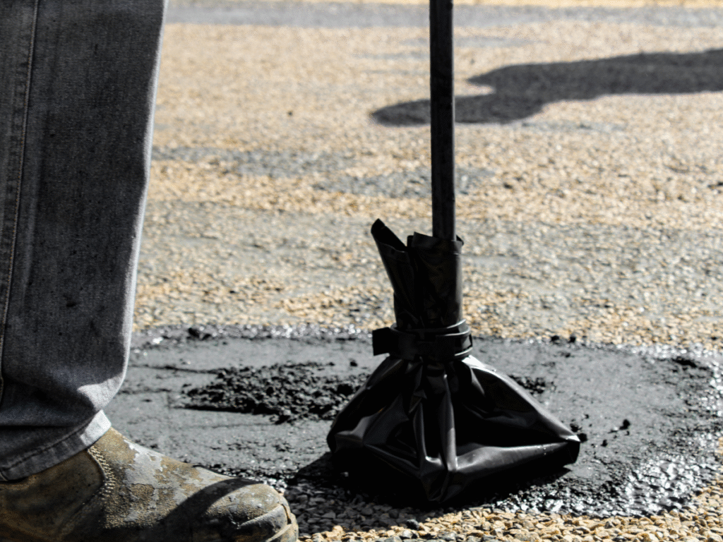 A person stands next to a tamping tool with a black plastic cover, compacting freshly poured asphalt on a road—showcasing the expertise of an Ohio paving and construction professional. The lower part of their jeans and work boot are visible.