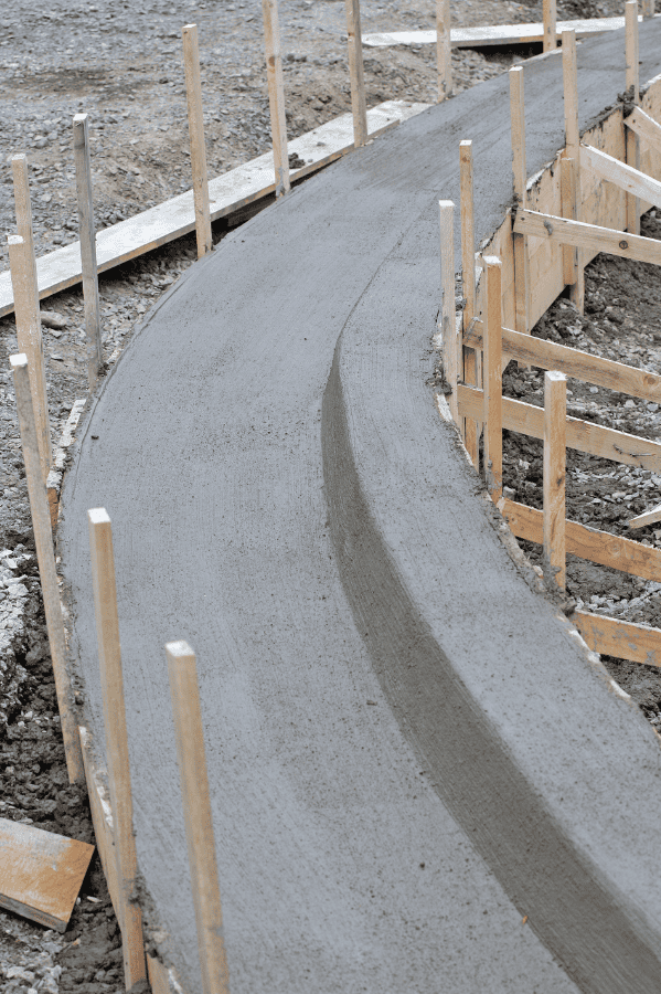 Freshly poured concrete forms a curved path or curb, supported by wooden stakes and formwork on a construction site with gravel and dirt—quality results you expect from top commercial asphalt companies near me.