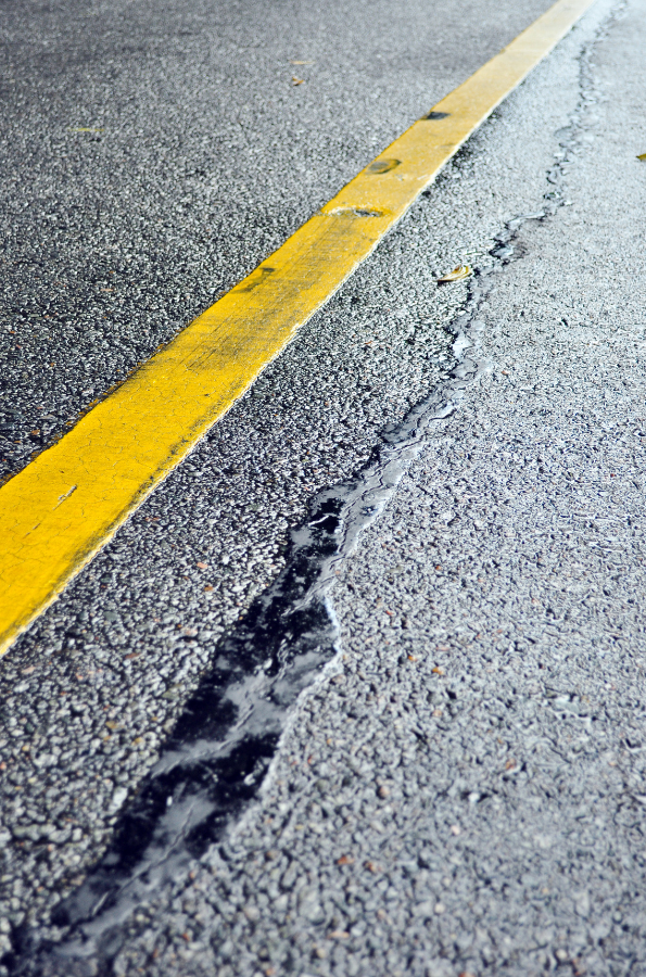 A close-up view of a cracked asphalt road with a yellow line running diagonally, and a visible black crack filled with sealant beside the line—showcasing the expert repairs done by asphalt companies Cleveland Ohio trusts.
