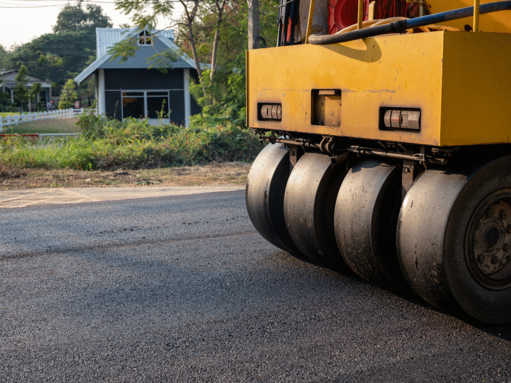 A yellow road roller, from one of the top asphalt companies Cleveland Ohio offers, compacts fresh asphalt on a road. A small house and greenery sit in the background, with the focus on the machine’s large black rollers.