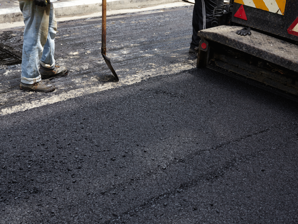 Two workers are resurfacing a road with fresh asphalt. One holds a shovel near the newly laid pavement, while part of a commercial asphalt companies near me machine is visible. The workers legs and textured asphalt surface are in focus.