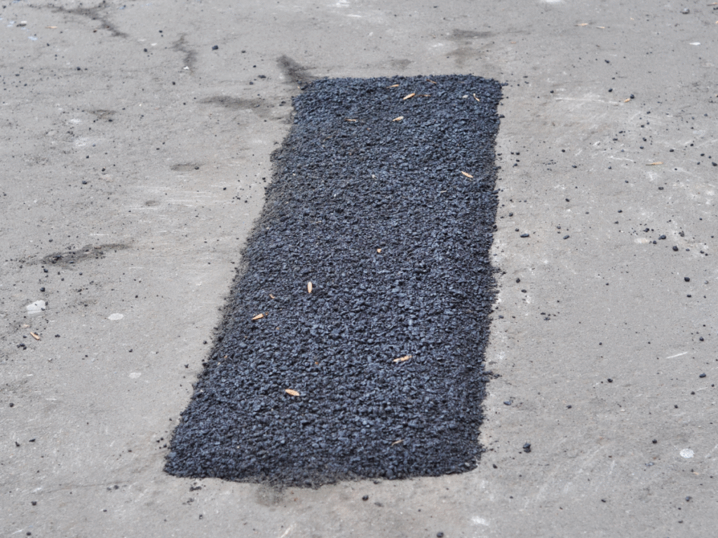 A rectangular patch of fresh black asphalt, laid by a leading commercial concrete paving contractor, covers a section of gray concrete pavement. The new asphalt stands out due to its darker color and rougher texture compared to the surrounding surface.
