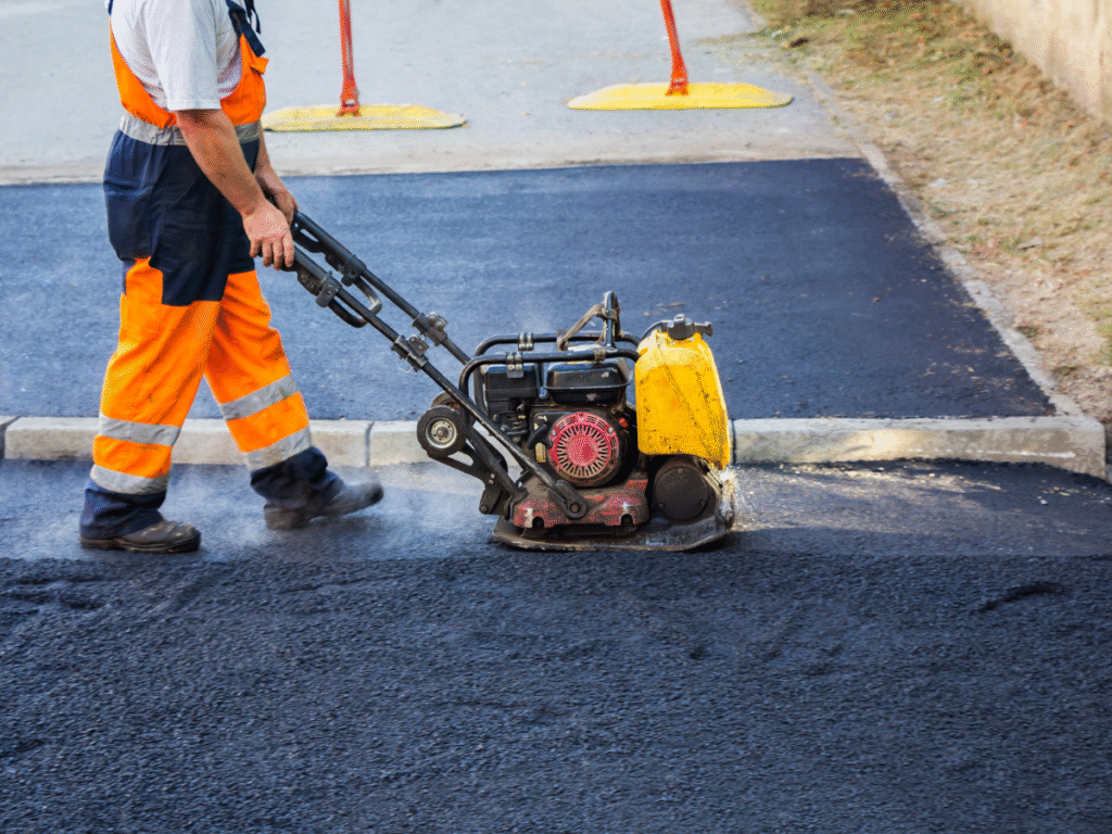 A construction worker in orange pants uses a yellow compactor machine to flatten fresh asphalt on a road. Safety barriers are in the background, typical of ohio paving and construction or commercial asphalt companies near me.