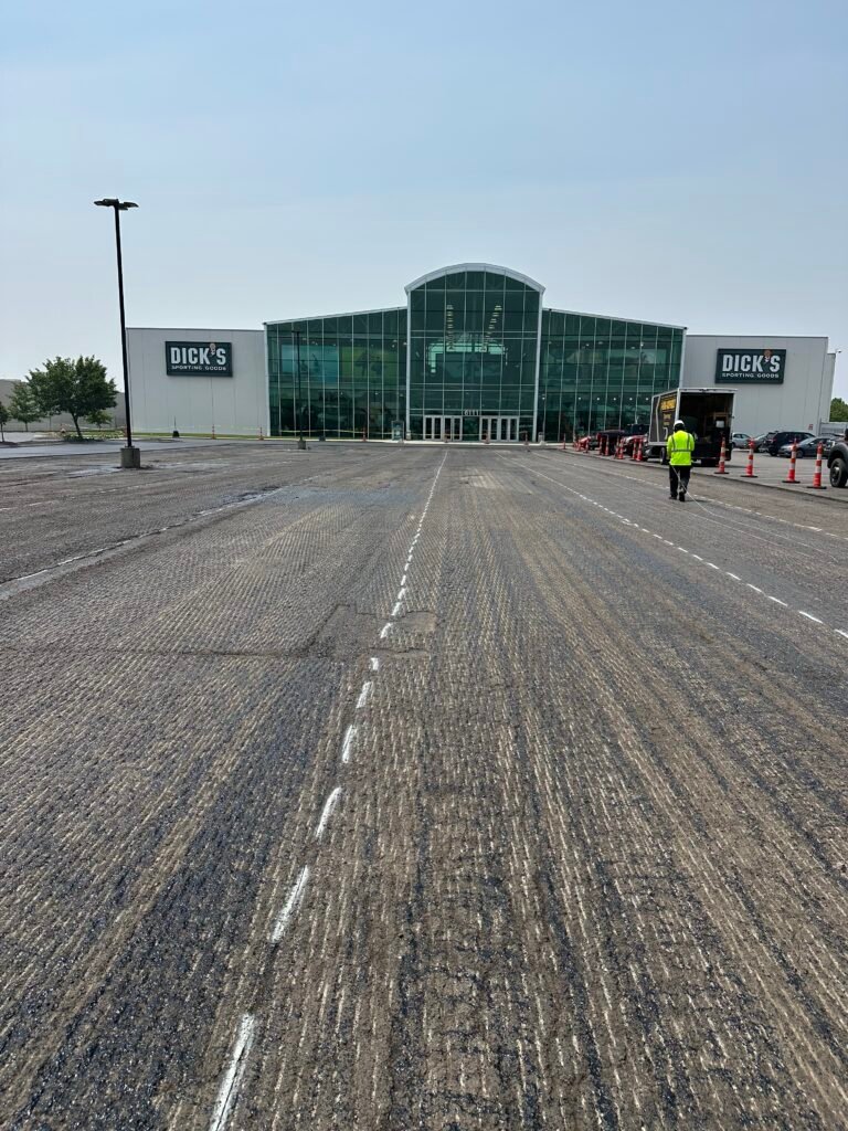 A large Dicks Sporting Goods store with a glass entrance is seen in the background. In the foreground, a rough, partially repaved parking lot stretches ahead, with a worker in a yellow vest and orange cones on the right.