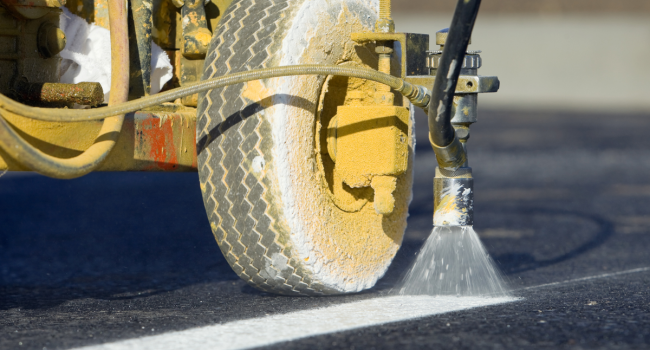 1 Close-up of a road marking machine’s wheel and nozzle spraying white paint onto asphalt, showcasing the precision work of Ohio Paving and Construction to create a straight line on the road surface.