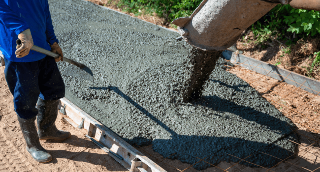1 A worker wearing boots and gloves stands next to wet concrete being poured by a commercial concrete paving contractor onto prepared ground, shovel in hand and greenery in the background.