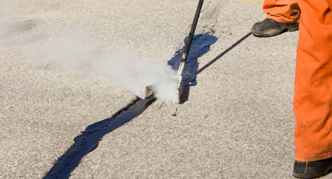2 A worker in orange pants repairs a crack in the road with hot sealant for one of the top asphalt companies Cleveland Ohio offers. Only the worker’s lower legs are visible, with steam rising and yellow road lines in the background.