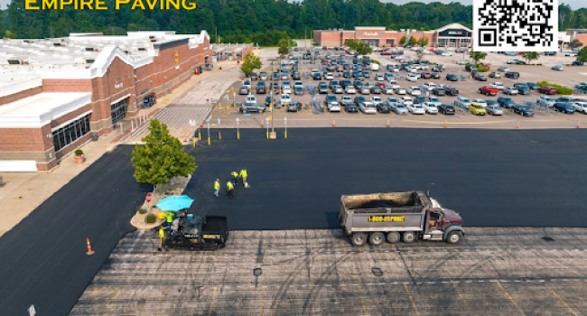 2024-11-12 Aerial view of a parking lot being paved by Empire Paving’s skilled team and machinery near a shopping center, with a busy parking area in the background. “Empire Paving” text and QR code appear in the top left corner.
