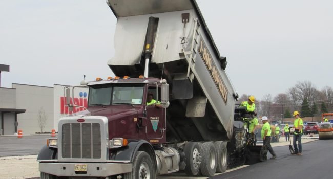 491439020_17858565234402229_3183972665482341584_n A dump truck unloads asphalt into a paving machine as several construction workers in safety gear, part of a leading commercial asphalt company near me, guide the process and work on freshly laid pavement by the roadside.