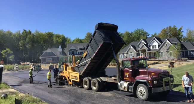 70677014_723767684716212_4819516805821235200_n A construction crew from Empire Paving is paving a residential street with an asphalt paver fed by a raised dump truck. Workers in safety vests are present, with houses and trees in the background under a clear blue sky.