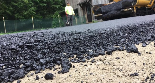 86733859_851599825266330_6237266797058326528_n Close-up of fresh asphalt being laid over a dirt road by a yellow paving machine from Ohio Paving and Construction, with a worker in a neon yellow shirt nearby and a wooden building and trees in the background.