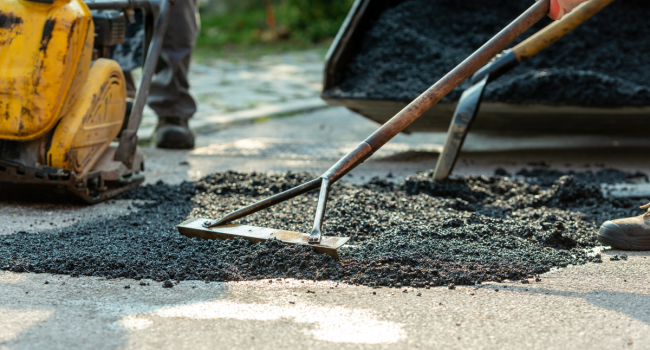 Close-up of a worker spreading hot asphalt on a road surface, with construction equipment and another worker in the background—showcasing the expertise of Ohio paving and construction professionals.