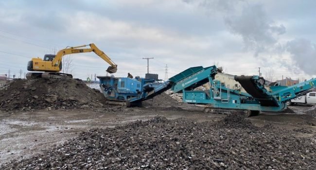 EMPIRE-MATERIALS-YARD A yellow excavator loads gravel into a blue and turquoise rock crusher, part of an empire paving operation at an outdoor construction site, with cloudy skies and scattered piles of stones in the foreground.