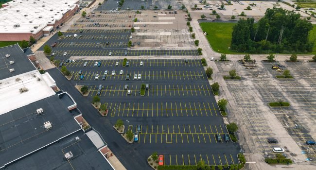 Empire-Paving-Aurora-Ohio-Wal-Mart-08-2023-8 Aerial view of a large, mostly empty shopping mall parking lot with a few scattered cars, surrounded by trees and commercial buildings—expertly maintained by Ohio paving and construction professionals.