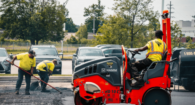 Rectangle-8 Three workers in yellow vests repave a parking lot; two use tools to spread asphalt while one operates an orange steamroller. Empire Paving, a trusted commercial asphalt company near me, handles the project as cars and trees are visible in the background.