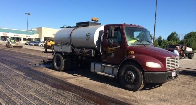 Resurface-5-800×600-1 A maroon and silver tanker truck from Empire Paving applies liquid to a parking lot during daylight, likely for paving or sealing, with a shopping center and several vehicles in the background.