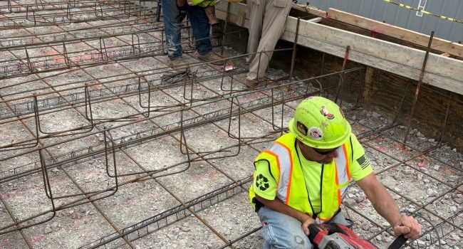 Construction workers in safety vests and helmets work on a rebar framework at a building site. One worker uses a power tool on the ground while others measure or inspect near a wooden form. Safety cones and tape are visible.