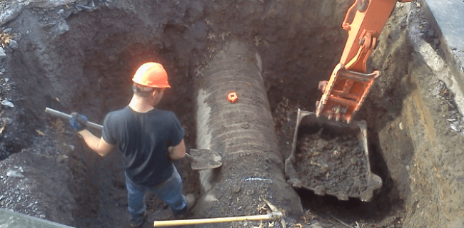 sewer-work-image-632×477-1 A construction worker in a hard hat stands in a deep hole beside a large, partially buried pipe while an orange excavator scoops dirt nearby, showcasing the expertise often found with commercial asphalt companies near me like Empire Paving.